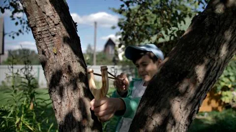 Boy hides behind a tree and aims at someone with a slingshot Stock Photos