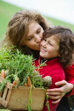 Boy holding a crate of vegetables with his mother kissing him Stock Photos