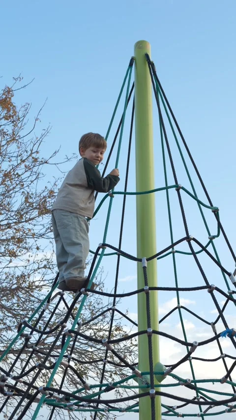 Boy holding rope net at the top of playground structure, vertical outdoor play Video stock 329277504
