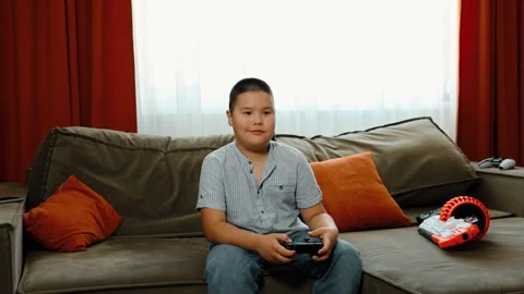 Boy holds game controller on sofa, striped shirt and jeans, video game play Video stock 312661491