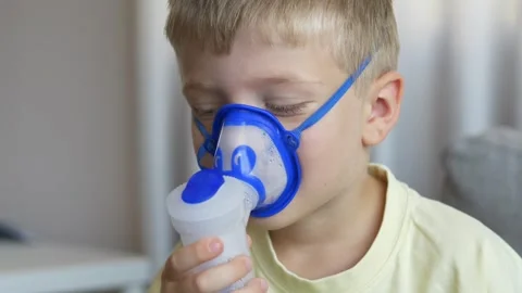 Boy holds nebulizer device himself while breathing through mask looking down in Stock Footage 345654300