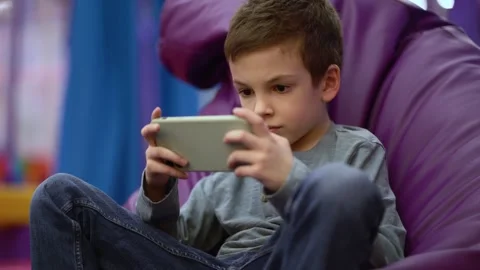 Boy intently plays a game on his smartphone, reclining in a large purple bag Stock-Footage 337199246