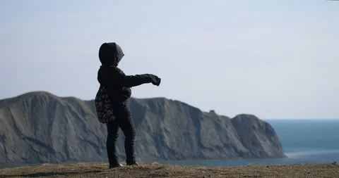 A boy in a jacket with deflated arms is standing on a hill by the sea Stock Footage 119563759