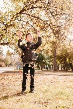 A boy in a jacket scatters leaves in the autumn park. The child rejoices at t Stock Photos