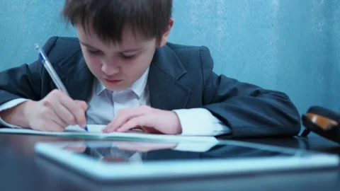 A boy in a jacket writes in a notebook with a pen at the table. in front of him Stock Footage 144179790