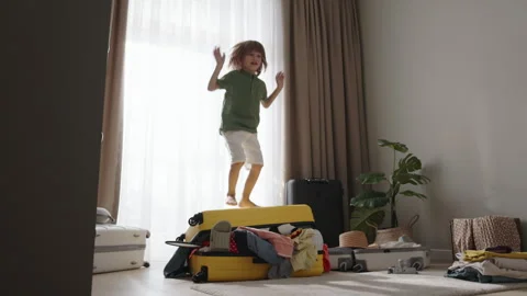 Boy joyfully jumps on packed suitcase. Child happily jumps on chaotically messy Stock-Footage 252530009