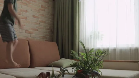 A boy jumping on sofa with dinosaur figurines on the table. The little explor Stock Footage 147673601