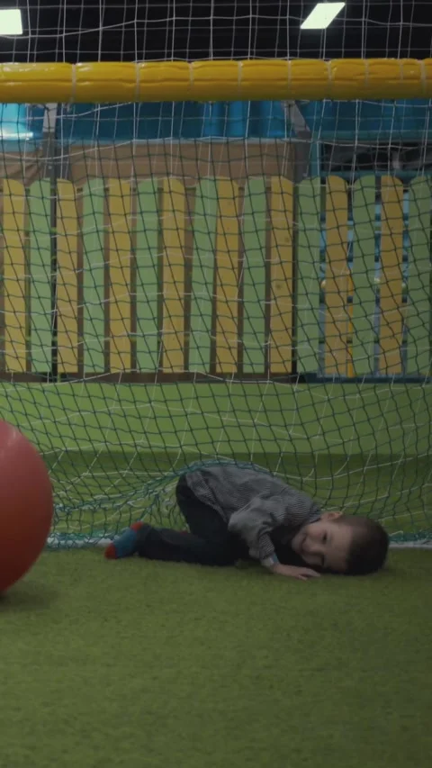 Boy kicking large red ball behind green safety net in colorful indoor playground Vídeos de archivo 342296534