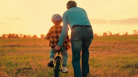 Boy kid parent dad play together, sunset. Child dreams to learn to ride bike 스톡 동영상 316607472