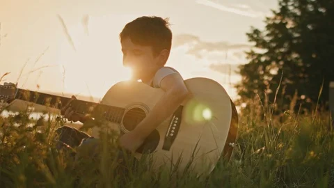 Boy kid in summer field training to play the guitar at summer sunset meadow Stock Footage 87502024