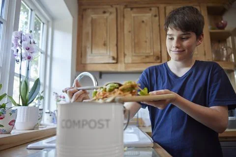 Boy In Kitchen Making Compost Scraping Vegetable Leftovers Into Bin Foto stock