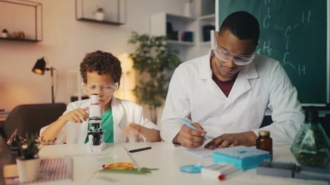 Boy in lab coat places sample under microscope, father documents test result Stock Footage 274836880
