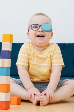 Boy in lazy eye patch laughs sitting in the children's room and plays with cubes Stock Photos