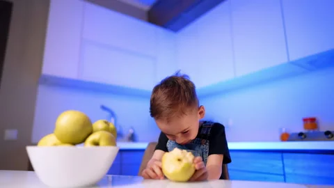 Boy leaning forward to eat yellow fruit at table. A child wearing denim overalls Stock Footage 343381330