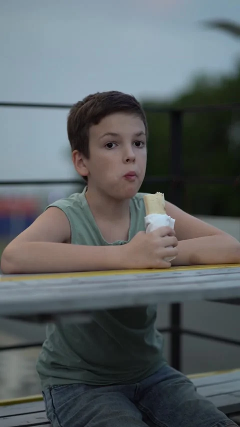 Boy leans on cafe table outdoors at dusk, eating a wrapped hot dog Stock Footage 341410006
