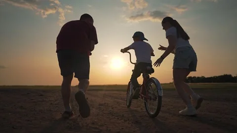Boy learns to ride with help from mother and father holding his bicycle. Family Stock Footage 318447709