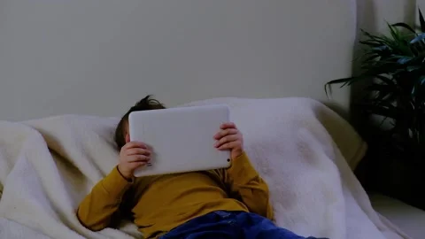 The boy lies on the bed and looks at something on the tablet.  Stock Footage