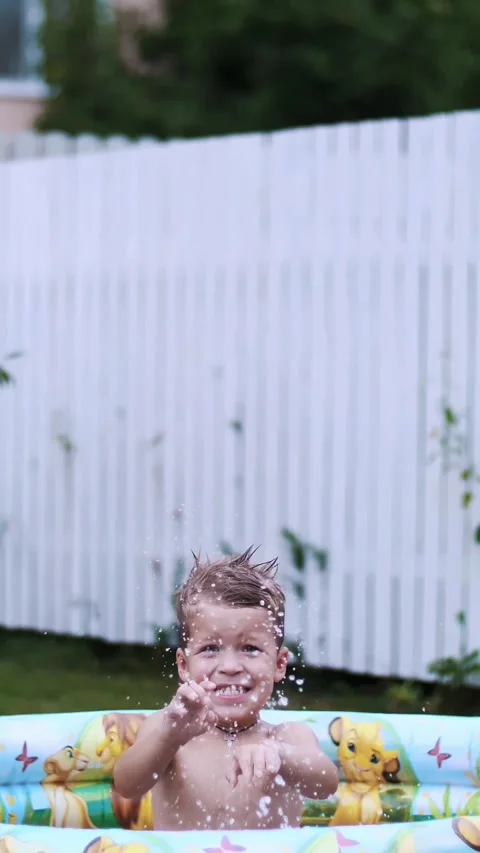 Boy lies in a children's inflatable pool, splashing water, smiling. Nearby float Stock Footage 311996467