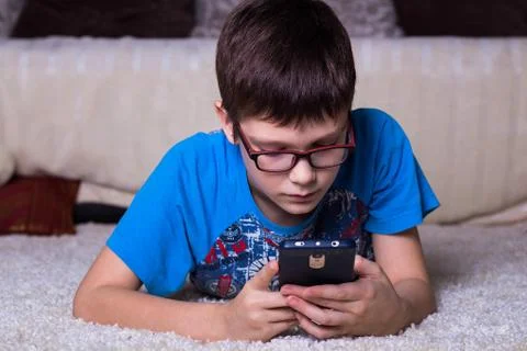 The boy lies on the floor facing the camera and looking at the smartphone Stock Photos
