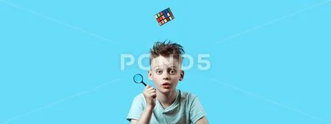 A boy in light t-shirt holds a small magnifying glass in his hand and a Rubik Stock Photos