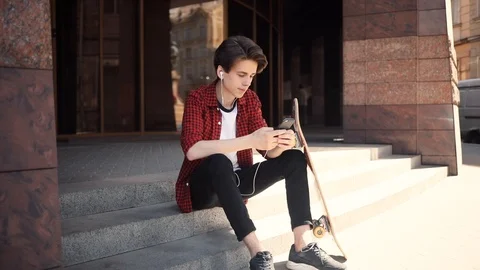 Boy look at camera using mobile phone with headphones keeps skate smile guy Stock Footage 92051646