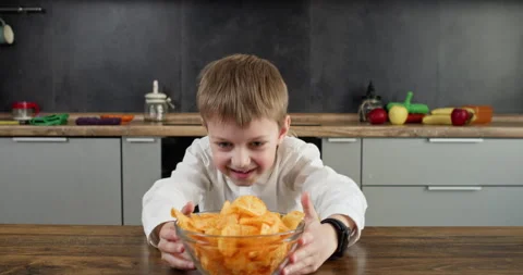 Boy looks down at potato chips with joyful smile pulling bowl Stockbeeldmateriaal 314720608