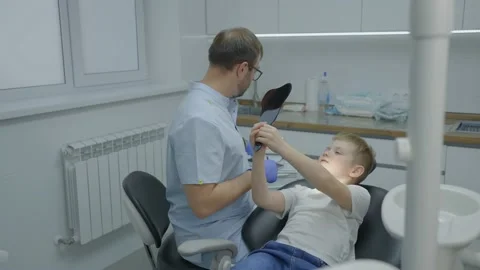 Boy looks in the mirror while sitting in dental chair at dentists appointment Stock Footage 224165974