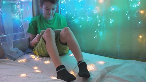 The boy looks at the tablet while sitting on a bed decorated with garlands Stock Footage 169357087