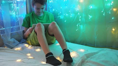 The boy looks at the tablet while sitting on a bed decorated with garlands Stock Footage 171129976
