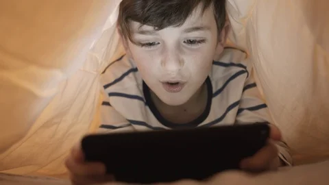 A boy is lying in bed under a yellow blanket playing into the game on the tablet Stock Footage 105688352