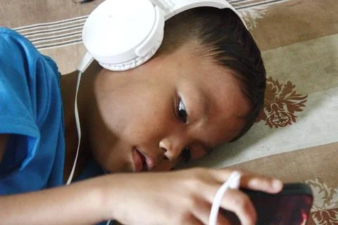 A boy is lying down playing with his cellphone using earphones. 스톡 사진