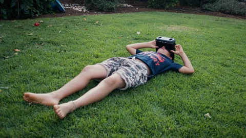 Boy lying on grass and using a virtual reality headset outdoors Stock Footage 275294660