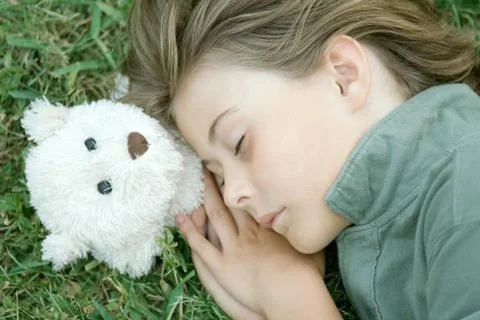 Boy lying in grass with eyes closed, head resting on teddy bear Stock Photos