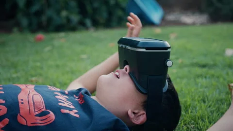 Boy lying on a grass lawn and moving his arms while using a virtual reality.. Stock Footage 275290443