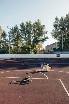 Boy lying on ground after fall from scooter on wide empty basketball court with Stock Photos
