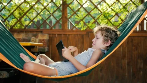 A boy lying in a hammock is playing on a tablet. The student works remotely in Stock Footage 205563747
