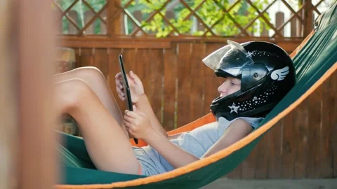 A boy lying in a hammock is playing on a tablet. A child in a motorcycle helmet Stock Footage 205563855
