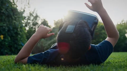 Boy lying on his back on grass and looking into a virtual reality headset Stock Footage 275288434