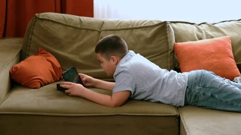 Boy lying on olive couch with digital tablet in blue striped shirt, indoor 動画素材 311506595