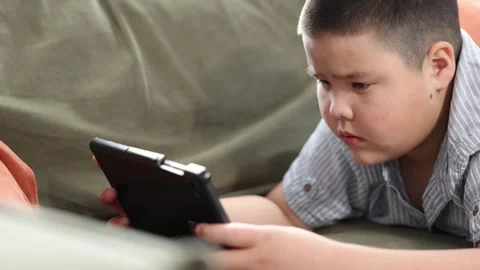 Boy lying on olive couch with digital tablet in blue striped shirt, indoor 動画素材 315429470