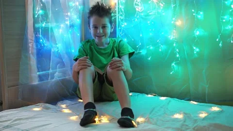 The boy is making faces sitting on a bed decorated with colorful garlands Stock Footage 171012464