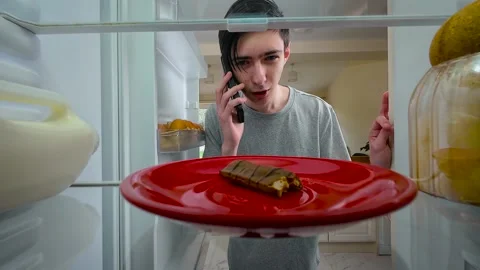 Boy opening fridge while talking on mobile phone, noticing bitten chocolate bar Stock Footage 328370573