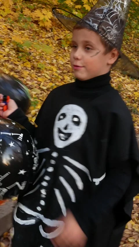 A boy with a painted skeleton walks with orange balloons in the autumn park Stock Footage 317849400
