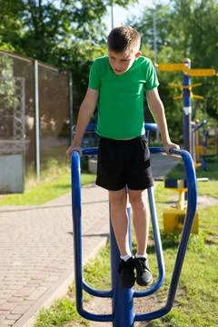 A boy performs dips on outdoor parallel bars. He's wearing a green shirt and  Stock Photos