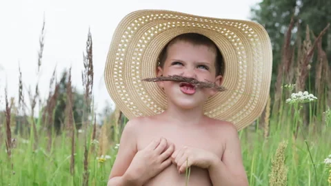 Boy playfully poses in straw hat, surrounded by lush greenery and wildflowers. Video stock 287691229