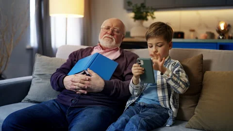Boy playing on cellphone while grandpa falls asleep with book, family moment Stock Footage 321358083