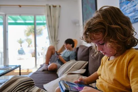 A boy playing on a digital tablet on the sofa at home, with his brother in the Stock Photos