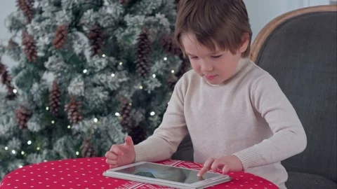 Boy playing games on his tablet sitting at the table near the Christmas tree Vídeos de archivo 70389532