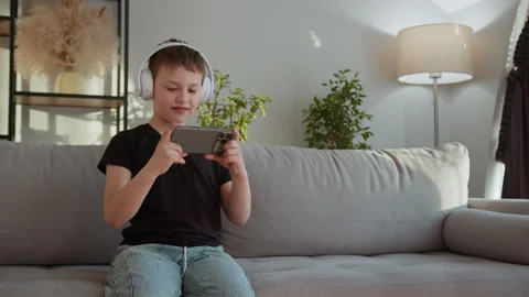 Boy Playing Games on Phone. Joyful child headphones mobile gaming cozy home Stock Footage 335478400