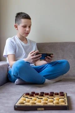 Boy Playing on Smartphone Instead of Checkers. A young boy in a white shirt and Stock Photos